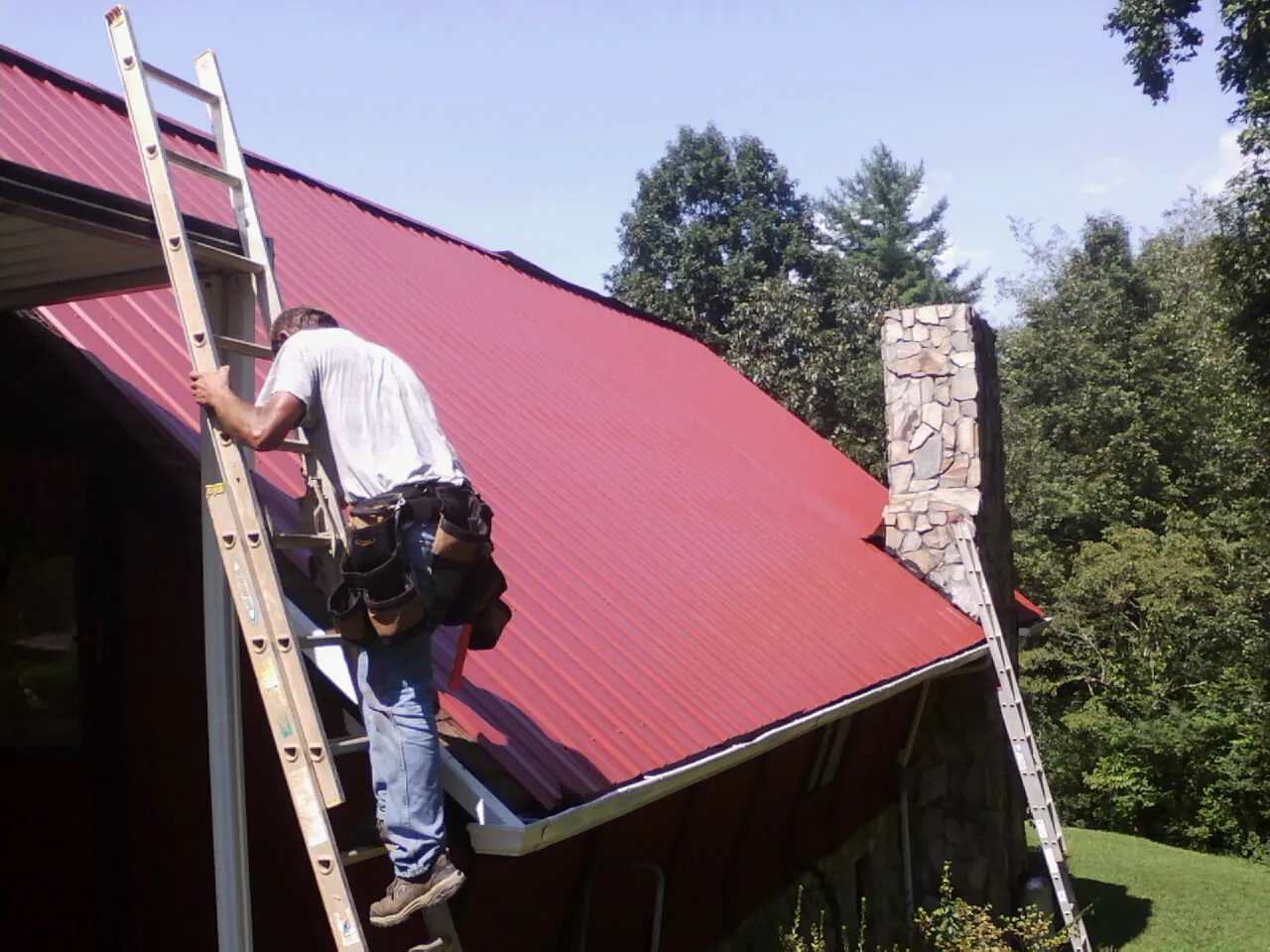 Roofer on a ladder beside a red metal roof and stone chimney — Soffit & Fascia Repair by Roofing Brothers in New Hyde Park
